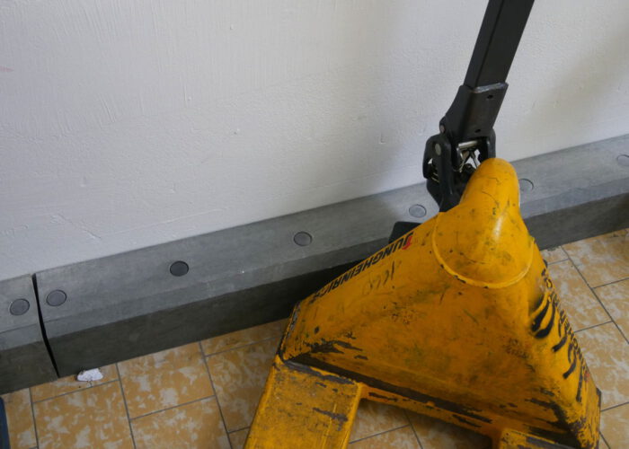 Ein Beispiel für Boden Rammschutz: Ein gelber Hubwagen, der von einer stabilen grauen Fußleiste gestoppt wurde, so dass die Wand nicht beschädigt wird.